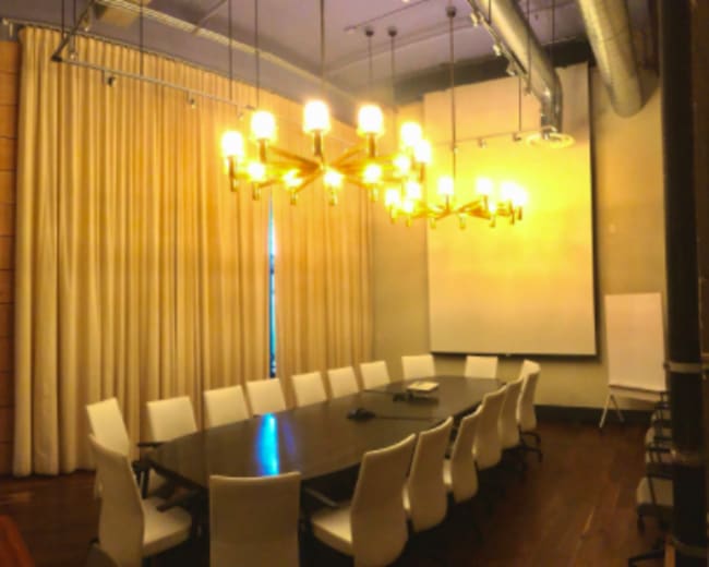 A conference room with a long wooden table and white chairs, illuminated by a chandelier, featuring large curtains and a projection screen.