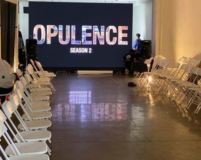 A spacious venue features white folding chairs arranged in two rows facing a large screen displaying "OPULENCE SEASON 2."