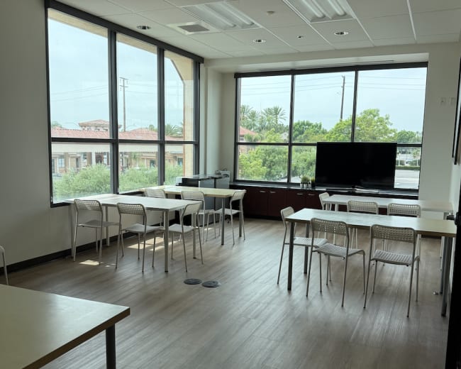 The image shows a spacious, well-lit meeting room with large windows, several tables and chairs, and a television facing the seating area.