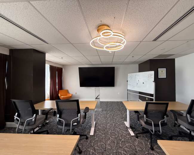 The image shows a modern conference room with multiple tables, chairs, a large screen on the wall, and a whiteboard.
