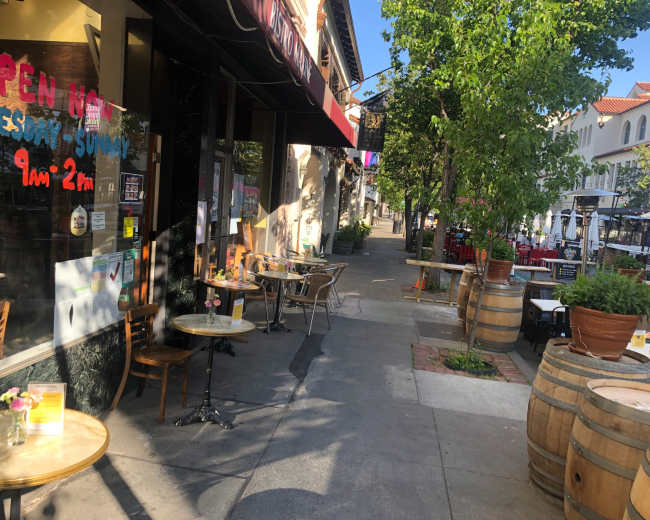 A sidewalk cafe features tables outside, with a storefront displaying an "Open" sign and nearby barrels lining the pathway.
