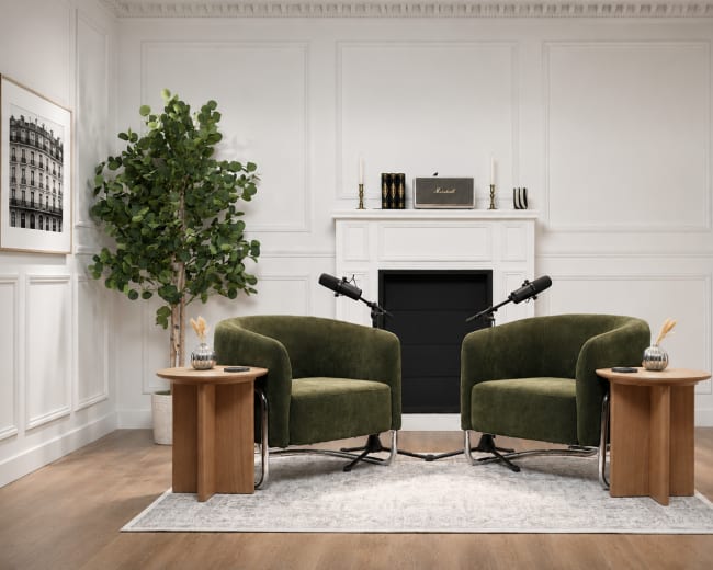 A minimalist interview setup features two green chairs facing each other with microphones on small wooden side tables, against a simple, elegant backdrop.
