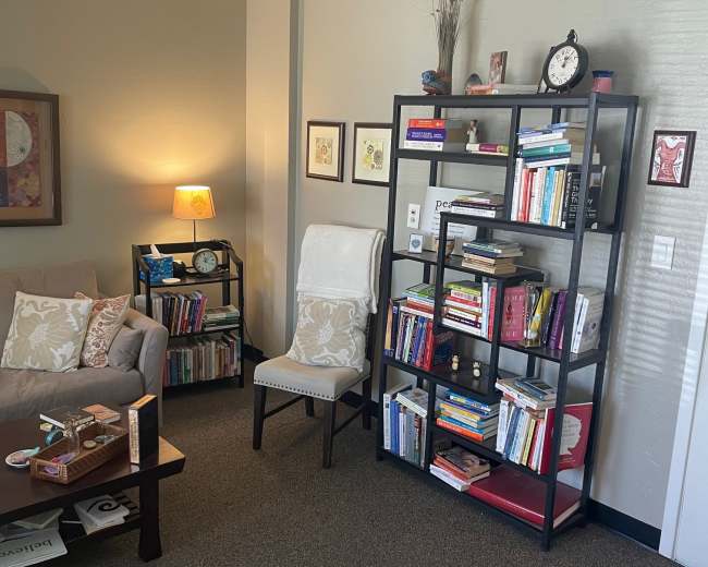The image shows a cozy reading area with a sofa, a bookshelf filled with various books, and a chair beside a lamp, all positioned in a well-lit room.