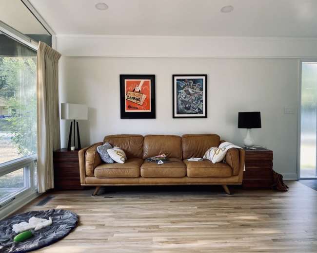 A living room featuring a brown leather sofa, two framed artworks on the wall, a side table with a lamp, and a wooden cabinet against the wall.