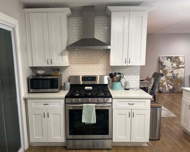The image shows a modern kitchen with white cabinets, a stainless steel stove, a hood above the stove, and a microwave, all set against a tiled backsplash.