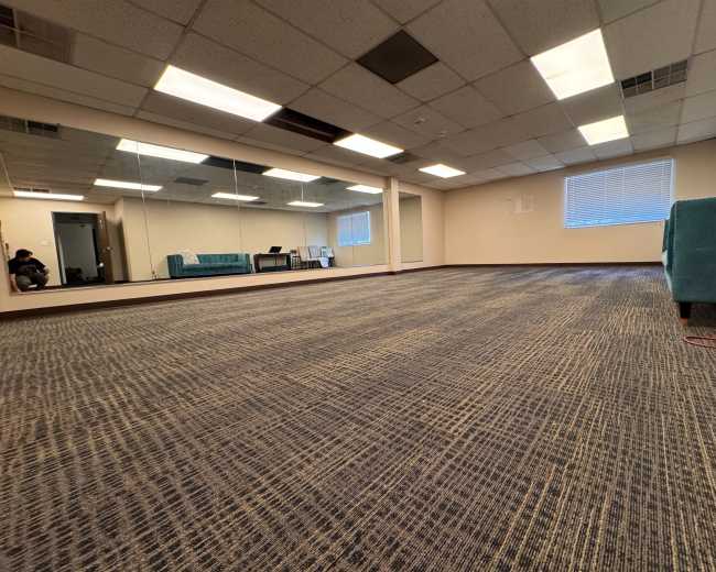 The image shows a spacious, empty room with carpeted flooring, a large mirror on one wall, and a few pieces of furniture in the corner.