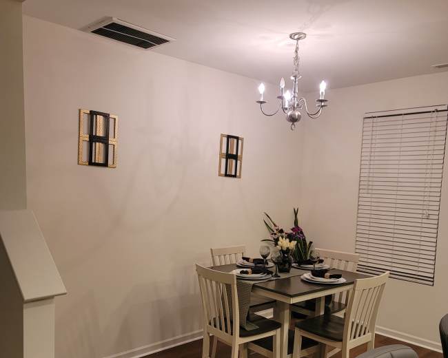 A dining area features a square table with four white chairs, a chandelier overhead, and two decorative wall pieces.