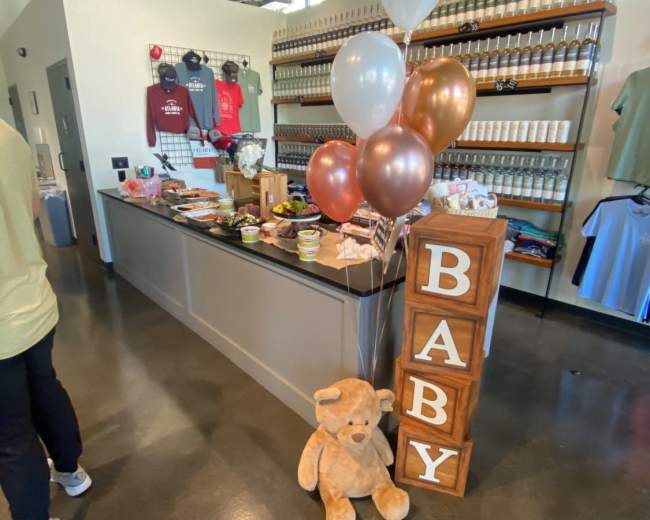 A table decorated with food and drinks is set up next to large balloons and a teddy bear, with a sign spelling "BABY" in block letters.