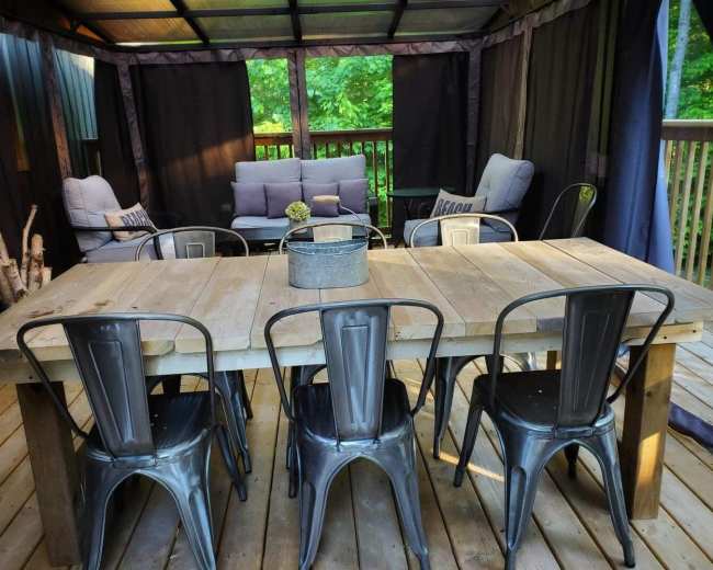 A wooden dining table with metal chairs is placed under a sheltered area surrounded by dark curtains and greenery.