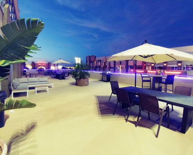 A rooftop patio features seating areas, tables, umbrellas, and decorative plants under a twilight sky.