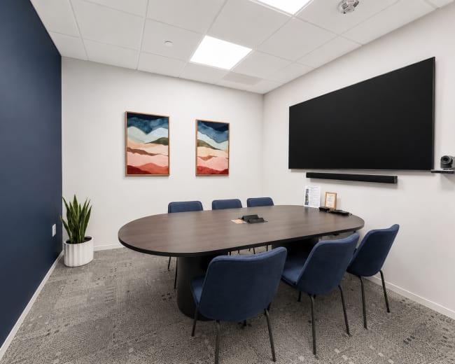 A modern conference room features an oval table surrounded by blue chairs, with two framed artwork pieces on the wall and a flat-screen monitor mounted above a shelf.