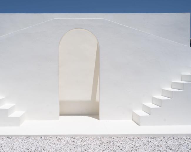 The image features a minimalist white architectural structure with an archway and stairs on either side against a clear blue sky.