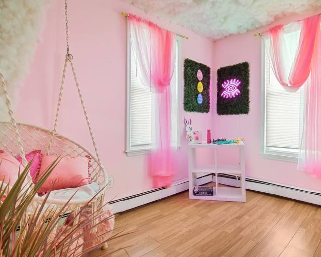 A pastel pink room featuring a hanging chair, sheer pink curtains, and decorative artwork on the walls.