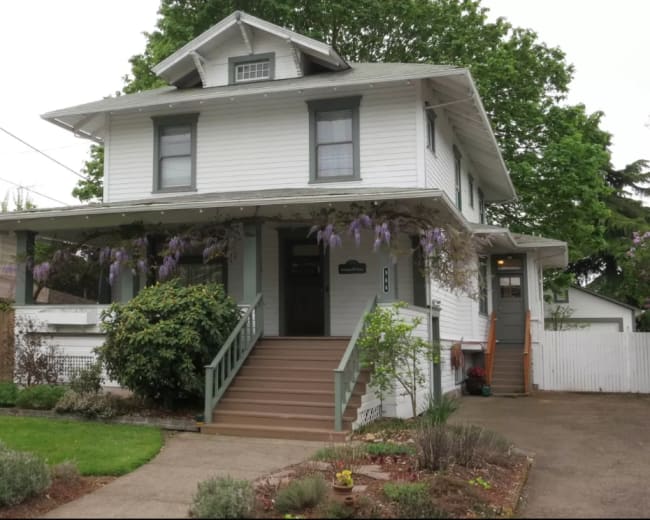 A three-story white house with a porch, surrounded by greenery and purple wisteria vines.
