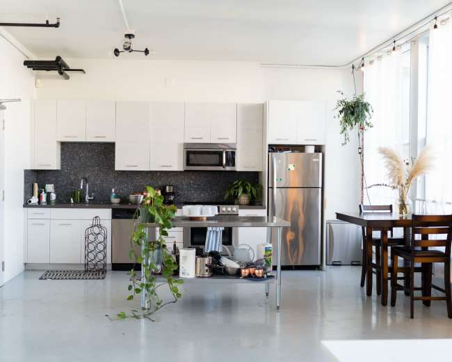 A modern kitchen and dining area features sleek cabinetry, a stainless steel refrigerator, and a table with chairs, accompanied by plants and decorative elements.
