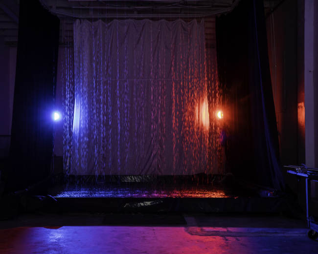 A curtain of water flows down from a suspended fabric backdrop, illuminated by red and blue lights in a darkened space.