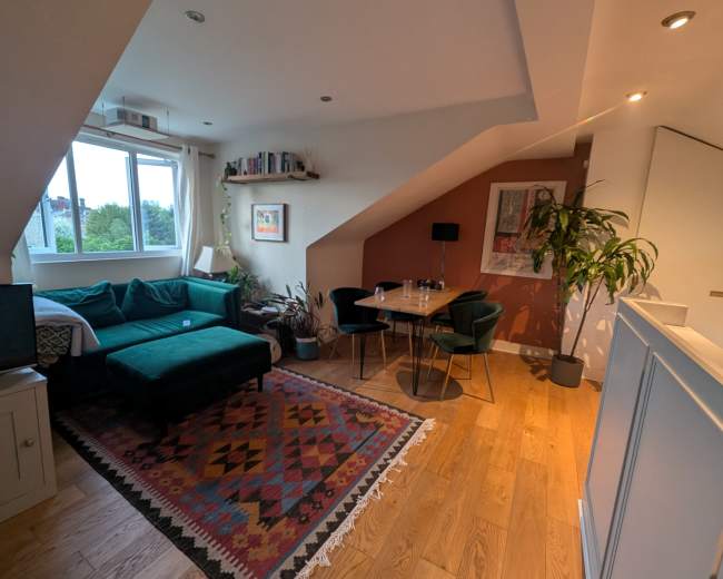 The image shows a cozy living room area with a green sectional sofa, a round dining table with chairs, and large windows providing natural light, set in a modern attic space.