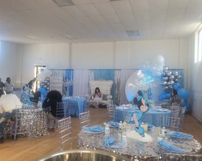 A decorated indoor venue set up for a celebration, featuring silver and blue decor, tables with sequined covers, and a large white throne chair.