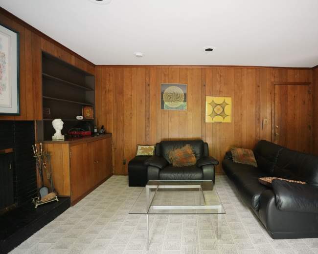 The image shows a living room with wooden panel walls, a black leather sofa, a glass coffee table, and a small decorative shelf.