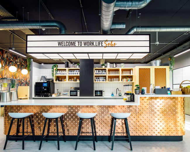 A modern kitchen area with a welcoming sign, a bar with stools, and shelves filled with various kitchen appliances and decor.