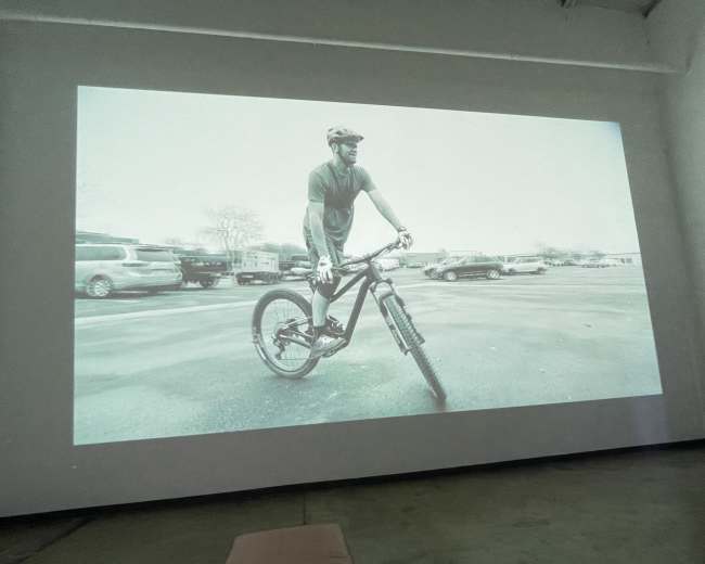 A black and white image of a man riding a mountain bike on one wheel, projected on a wall in a spacious room.