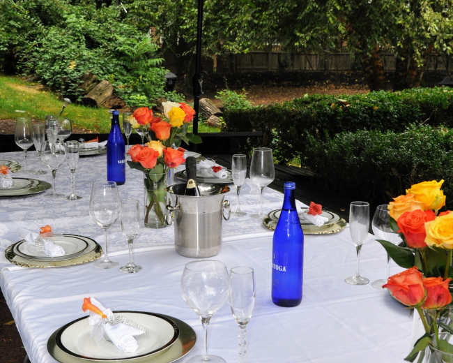 A dining table is set outdoors under a red umbrella, decorated with floral arrangements and accompanied by glasses and bottles of water.