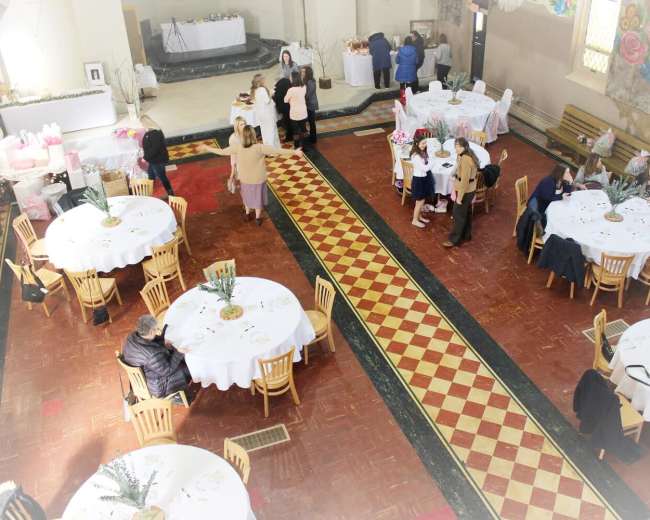 A spacious event hall is set up with round tables covered in white tablecloths, with guests mingling and a buffet area in the background.