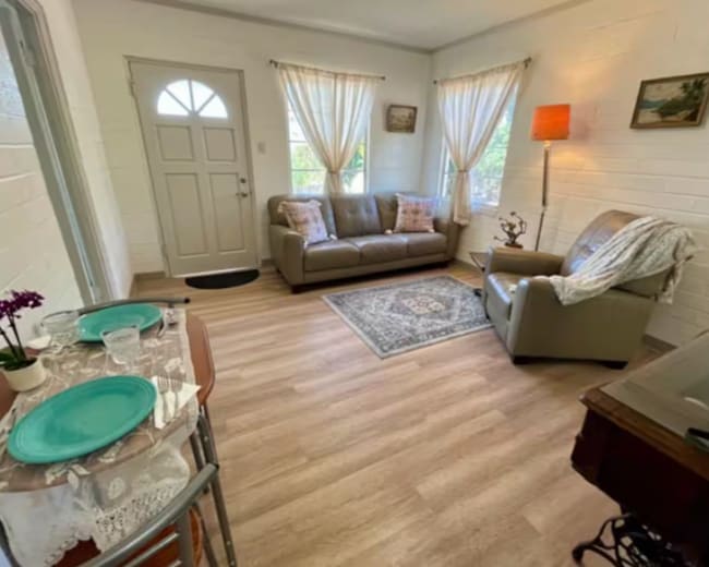 The image shows a small living room with a sofa, an armchair, a small dining table set for two, and a door leading outside.