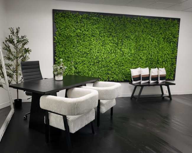 The image depicts a modern office space featuring a black table with two white upholstered chairs, a large mirror, a potted plant, and a green wall covered in artificial foliage.