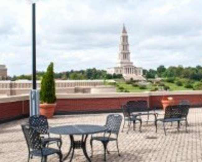 A rooftop terrace features several metal tables and chairs overlooking a landscaped area with a prominent building in the background.
