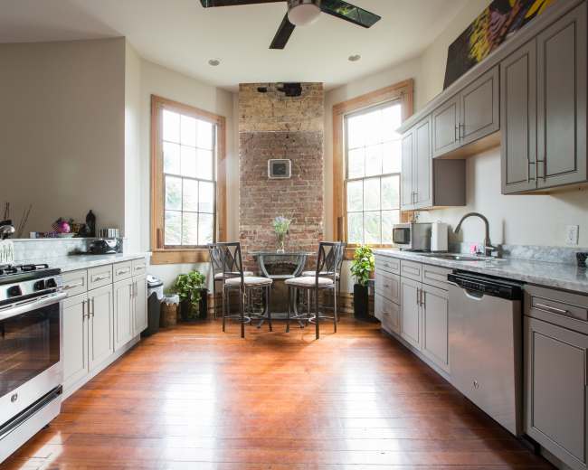 The kitchen features a combination of modern appliances and a rustic brick wall, with a small dining table set near large windows.