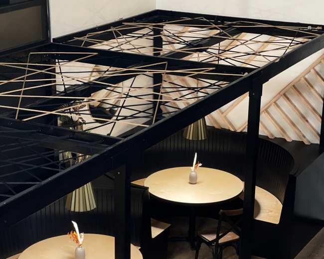 The image shows a modern dining area with two circular tables under a black lattice roof, each featuring a small vase and a candle as centerpieces.