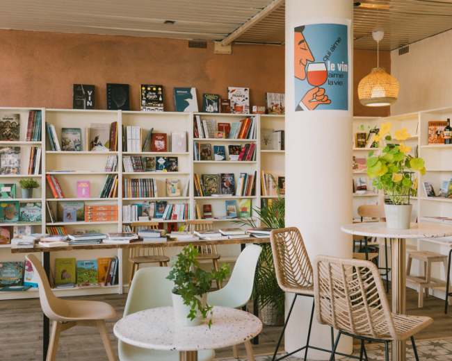 The image shows a bright and modern café-like space featuring bookshelves filled with various books and magazines, tables with plants, and decorative lighting.