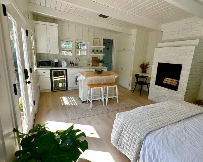 A modern, open-concept living space featuring a kitchen with white cabinets, a central island with seating, and a cozy area with a bed and a fireplace.