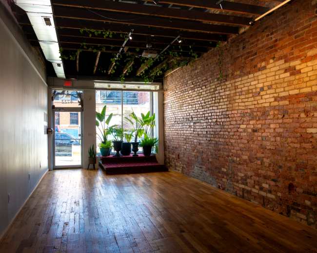 The image shows a spacious interior with exposed brick walls, wooden flooring, and potted plants near a large front window.