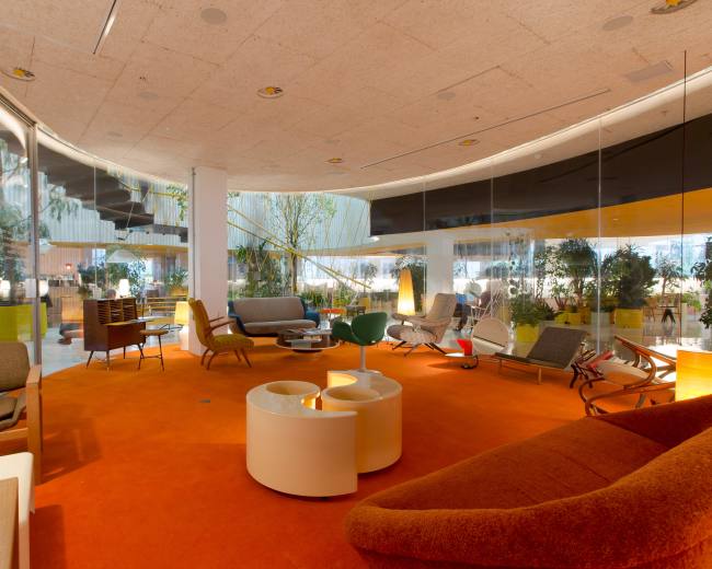 The image shows a modern lounge area with various seating arrangements, plants, and large windows overlooking an outdoor space.