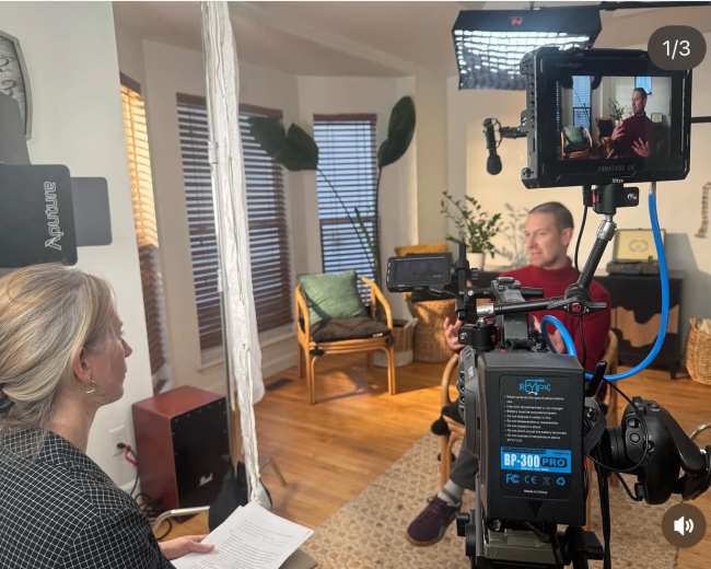 A man sits in a chair speaking to a woman who is positioned across from him, with a camera and lighting equipment set up for filming in a well-lit room.