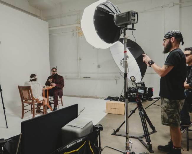 A photography studio is set up with a model sitting at a table while a photographer adjusts lighting equipment and other crew members assist.