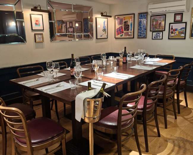 A wooden dining table is set for a meal in a well-decorated restaurant, surrounded by artwork on the walls and featuring chairs with burgundy cushions.