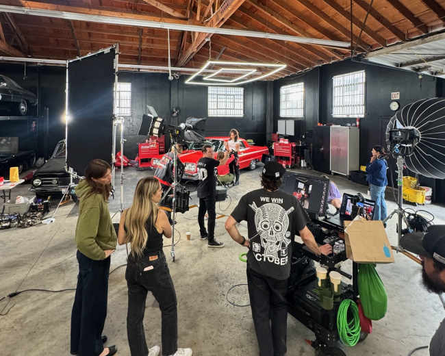 A group of people is working on a film or photo shoot in a spacious studio, with a red car positioned in the center under bright lights and equipment.