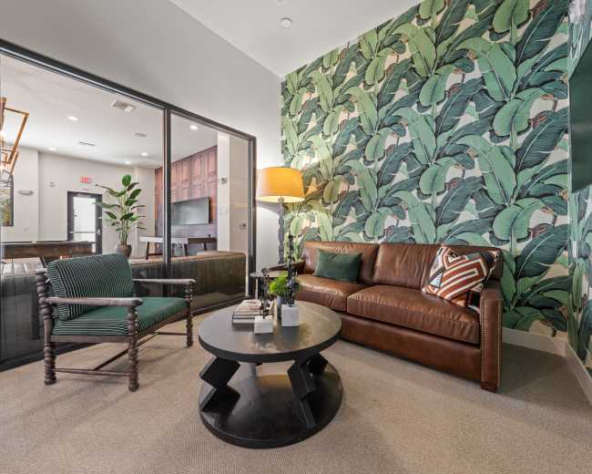 The image shows a cozy lounge area with a brown leather sofa, a green armchair, a round black coffee table, and a wall adorned with large banana leaf wallpaper.