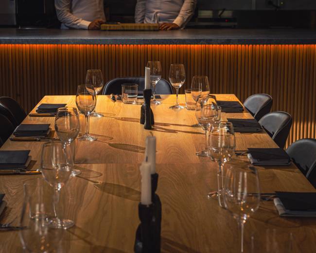 A large wooden dining table is set with glasses and black napkins, illuminated by warm light from the background.