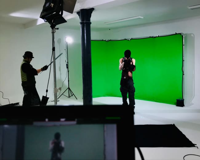 A person stands in front of a green screen while another individual prepares to shoot a video or film scene in a studio.