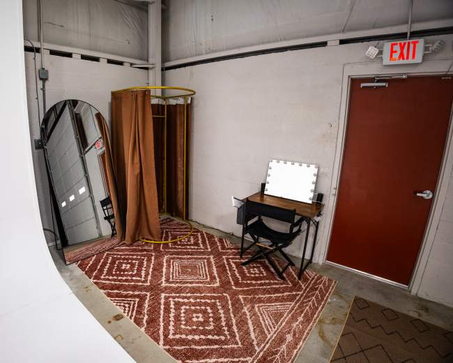The image shows a small, minimalistic room featuring a dressing area with a mirror, a brown curtain, a chair, and a lighted vanity table on a patterned rug.
