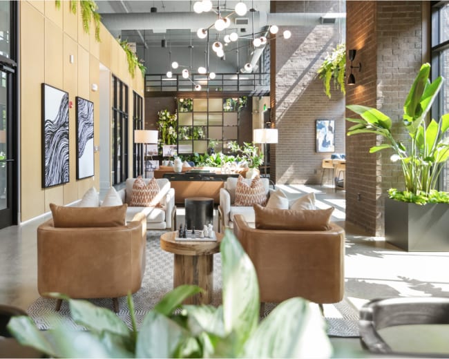 The image shows a modern lounge area featuring comfortable seating, decorative plants, and artwork against a backdrop of large windows and stylish architecture.