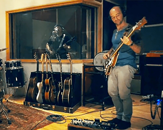 Two musicians are playing instruments in a recording studio, with one on drums and the other on guitar, while a vocalist is visible behind a glass panel.