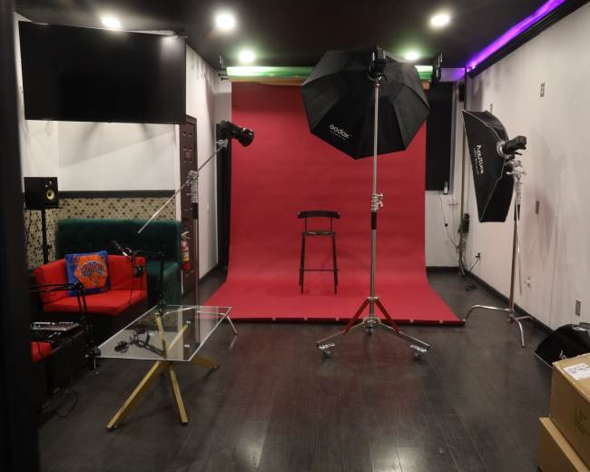A photography studio is set up with a red backdrop, a black chair, and lighting equipment, surrounded by a green couch and a glass table.