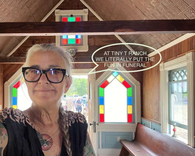 A woman stands inside a small, colorful chapel with stained glass windows, holding a phone and smiling at the camera.