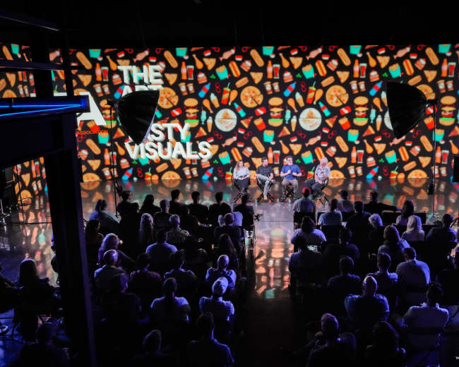 A group of speakers is seated on a stage in front of a large audience, with colorful visuals projected on the backdrop.