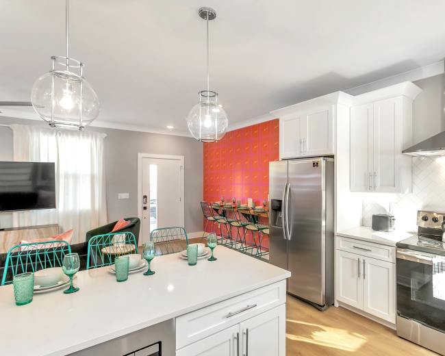 A modern kitchen features white cabinetry, stainless steel appliances, and a vibrant orange accent wall with a dining area visible in the background.